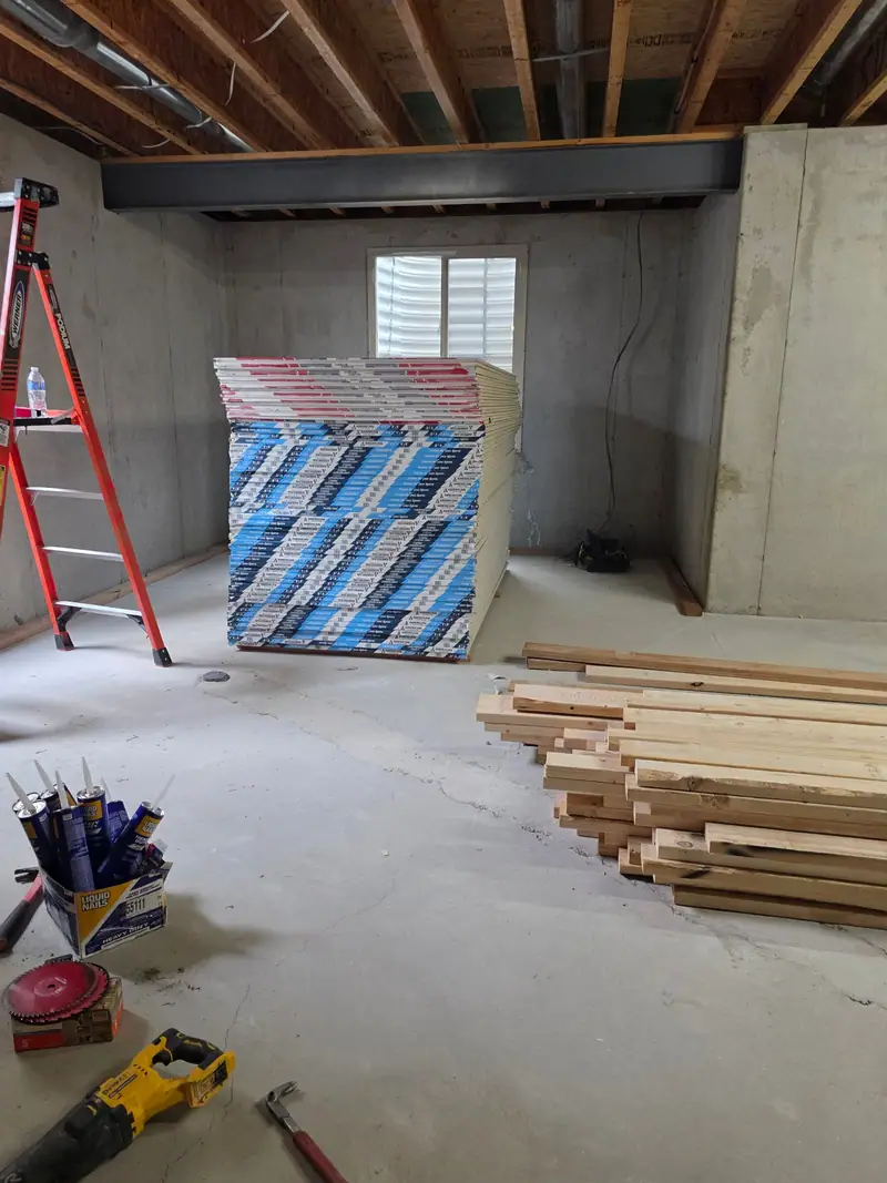 Drywall delivery stacked in an unfinished basement next to a ladder and lumber
