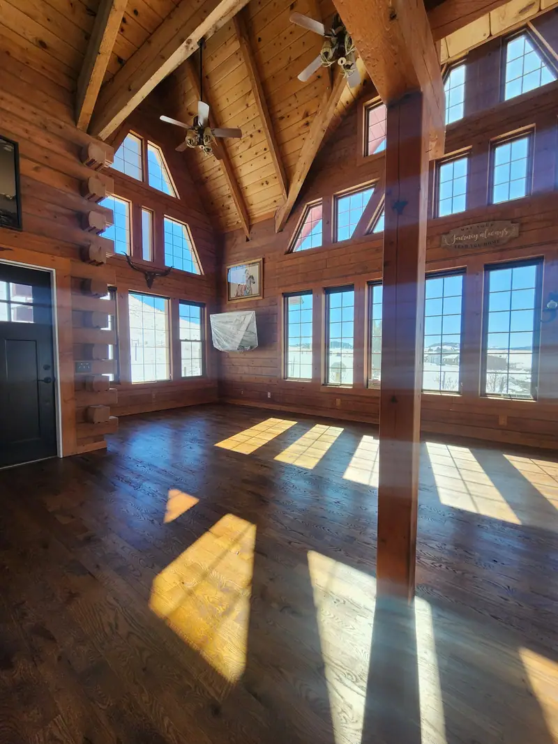 Log home great room with tall gable windows, timber trusses and sunlit hardwood floors