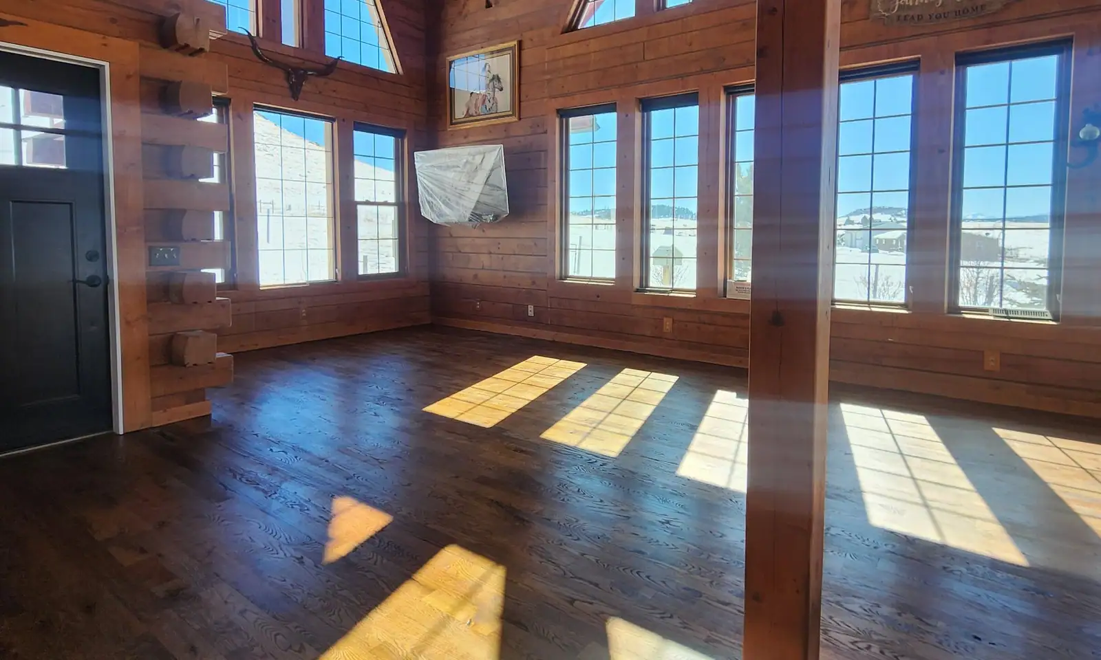 Log home great room with tall gable windows, timber trusses and sunlit hardwood floors