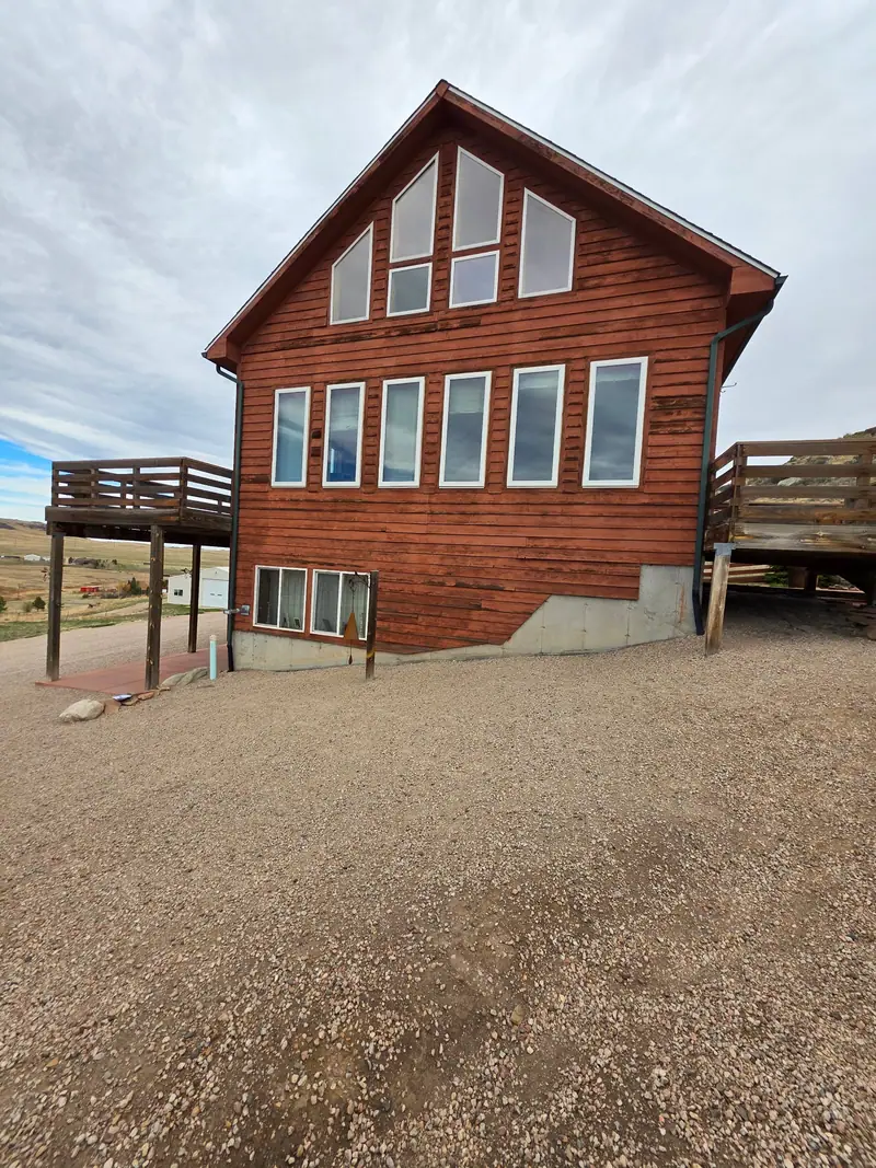 Wood-sided cabin with tall gable windows and decks on a gravel lot