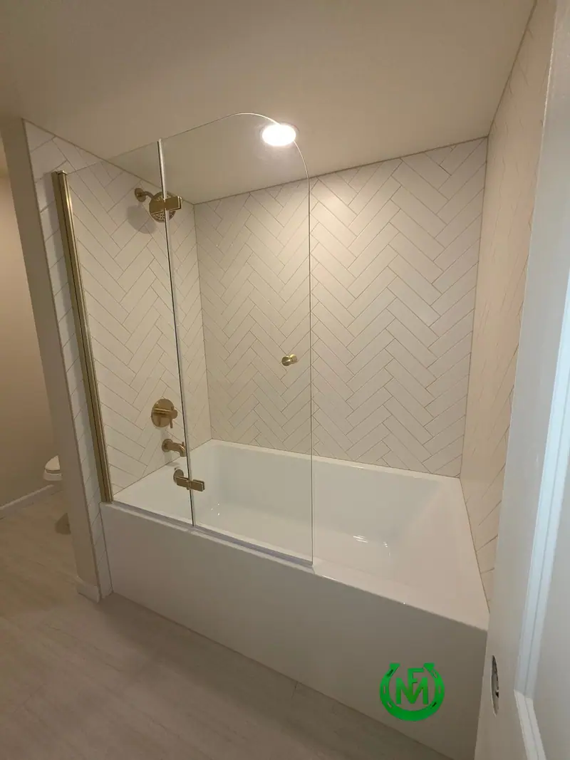 Tub and shower surround in white herringbone tile with brushed brass fixtures and a glass panel